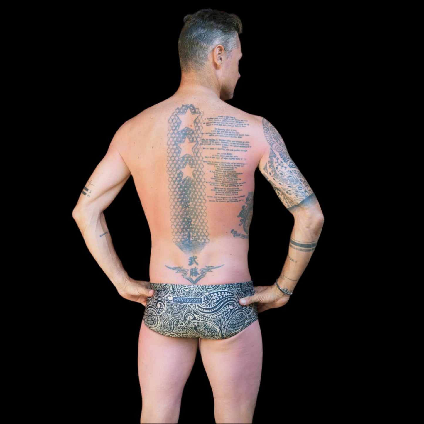 Fit man wearing gold and black Paisley pattern one piece swimwear with a brand logo hands on hips