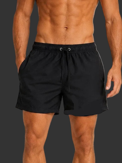A shirtless man modeling black swim trunks against a plain light gray studio background.