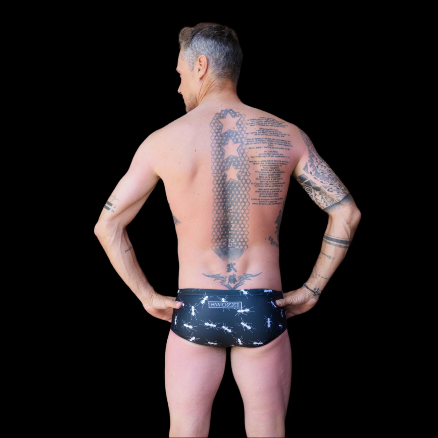 Fit man wearing black with white ants pattern one piece swimwear with a brand logo  hands on hips
