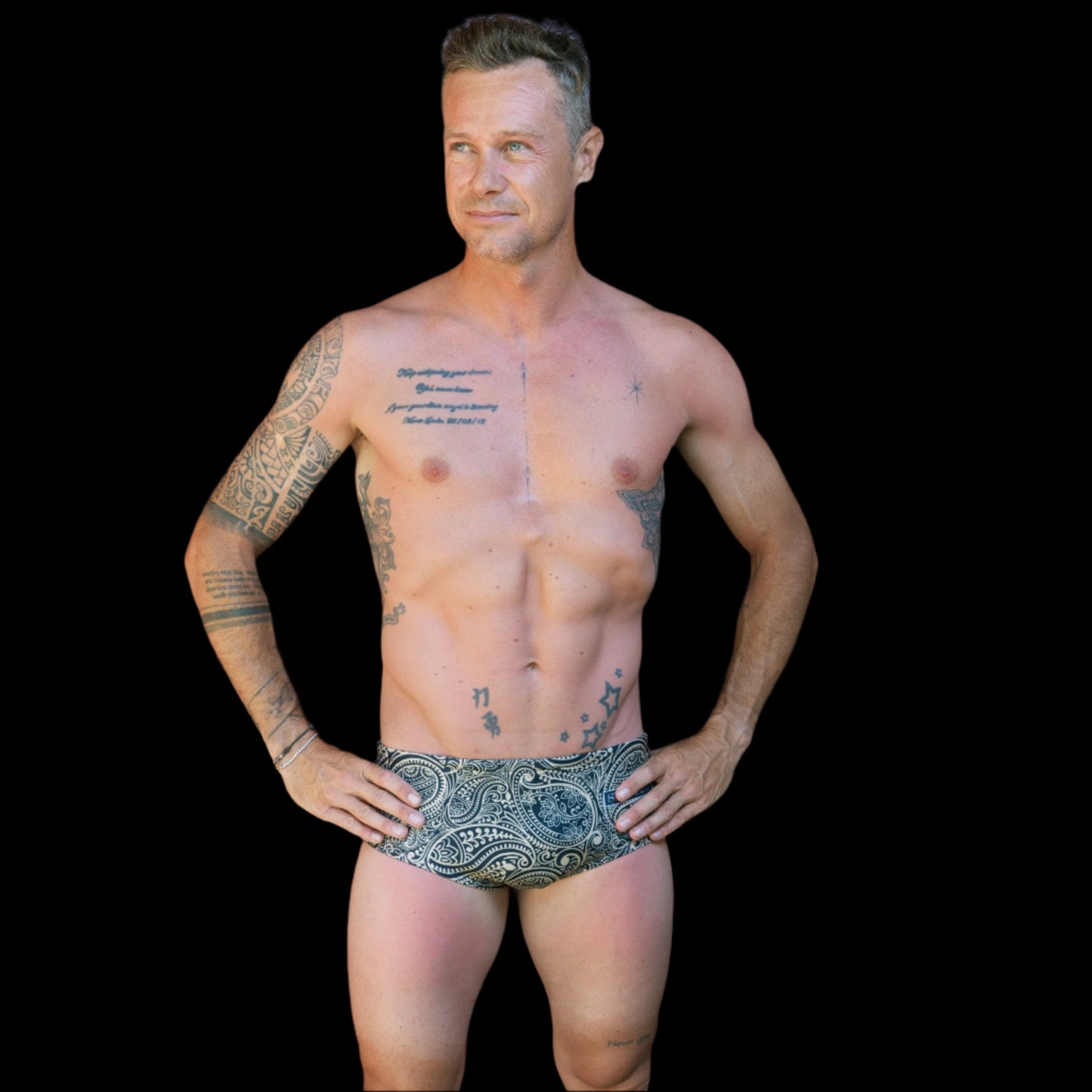 Fit man wearing gold and black Paisley pattern one piece swimwear with a brand logo   hands on hips